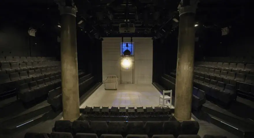Pusta scena w **teatrze stu** miejsc, z rzędami siedzeń po bokach. Na scenie łóżko, białe krzesło i reflektory.