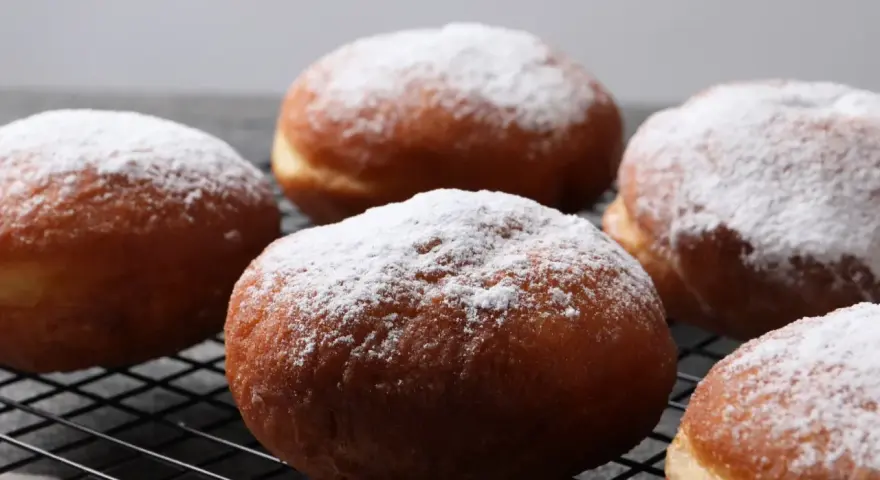 Pięć puszystych, pieczonych paczków, oprószonych cukrem pudrem, leży na czarnej kratce.