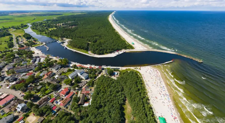 Widok z lotu ptaka na miasteczko nad zatoką, las i plażę. Na falochronie widać czerwoną latarnię morską kikut.
