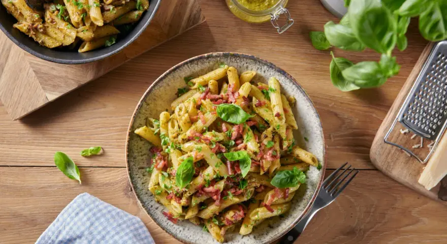 Makaron penne z pesto, boczkiem i bazylią na drewnianym stole.