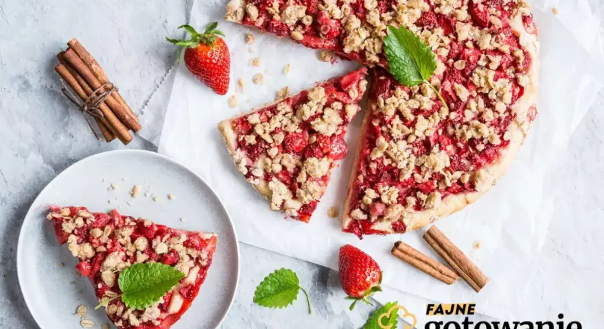 Pyszna tarta z truskawkami i kruszonką, udekorowana miętą, obok laski cynamonu i świeżych truskawek.