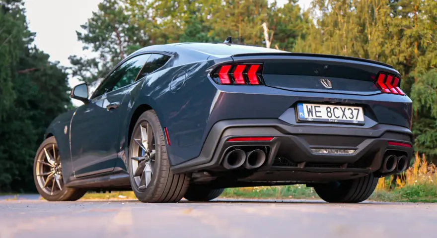 Ciemnoszary Ford Mustang, niczym wino dark horse, prezentuje swoje sportowe linie na tle zieleni. Widok z tyłu podkreśla mocne światła i podwójne końcówki wydechu.