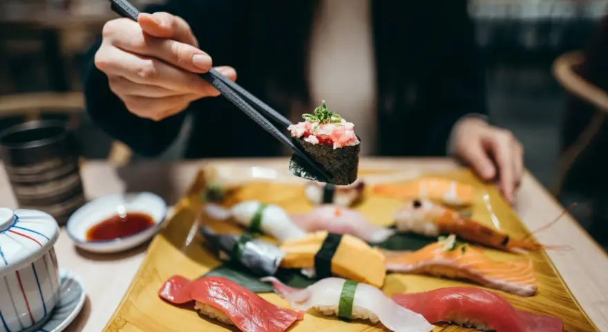 Eine Hand hält mit Stäbchen ein Stück Sushi mit gehacktem Thunfisch. Auf dem Teller liegen verschiedene Sushi-Rollen und Nigiri, bereit für den Genuss in Berlin.