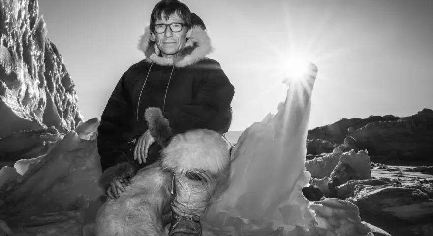 Ein Mann in traditioneller Inuit-Kleidung sitzt in eisiger Landschaft. Die Sonne scheint hell.
