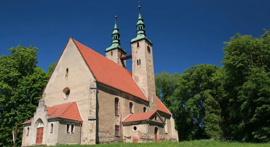 Stary kościół w stolcu z dwiema wieżami i zielonymi dachami, otoczony bujną zielenią drzew pod błękitnym niebem.