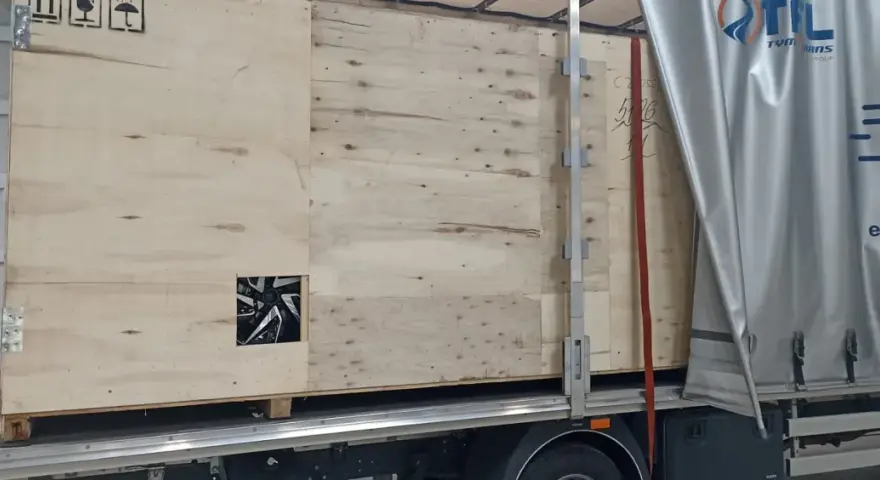 Drewniana skrzynia z widocznym fragmentem felgi, gotowa do transportu. Widać logo firmy transportowej i oznaczenia towaru.