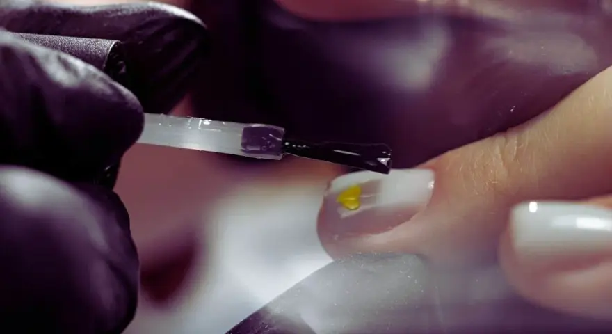Application d'un vernis transparent sur un ongle orné d'un petit cœur jaune. Le base coat, c'est quoi ? C'est la première étape pour une manucure parfaite.