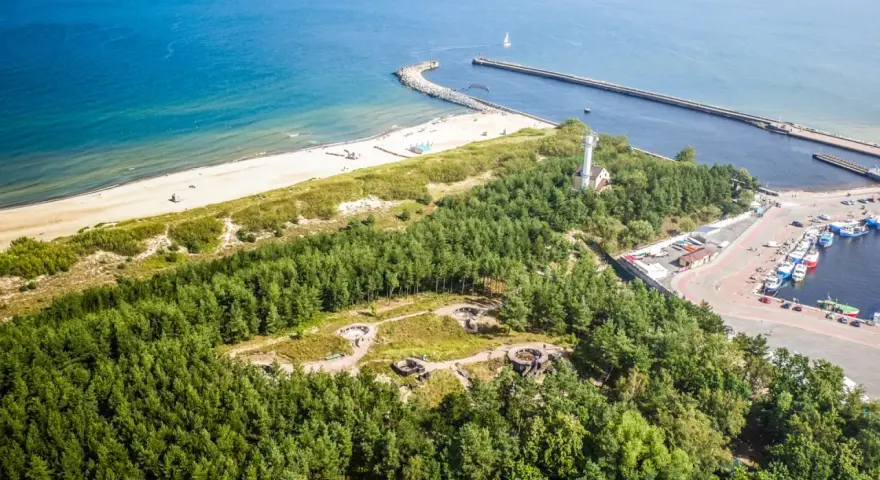 Widok z lotu ptaka na wybrzeże z piaszczystą plażą, lasem i portem. Wśród drzew widoczne są historyczne bunkry nad morzem.