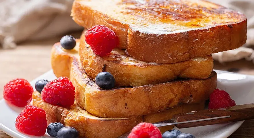 Una pila de tostadas francesas doradas, decoradas con frambuesas y arándanos, perfectas para aprender cómo se hacen las tostadas francesas.