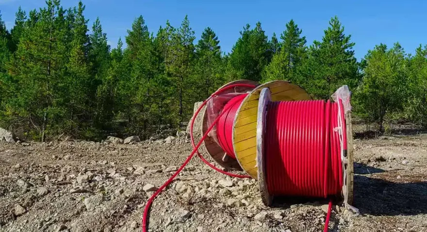 Czerwony kabel na drewnianym bębnie, przygotowany do uzbrojenia działki. W tle las i błękitne niebo.