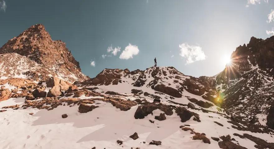 Ein Wanderer steht auf dem Gipfel des Jbel Toubkal, umgeben von schneebedeckten Felsen und einem strahlenden Sonnenschein.
