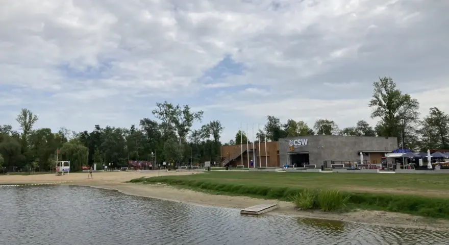 Spokojne jezioro małe żnińskie z piaszczystą plażą, budynkiem CSW i zielenią w tle.