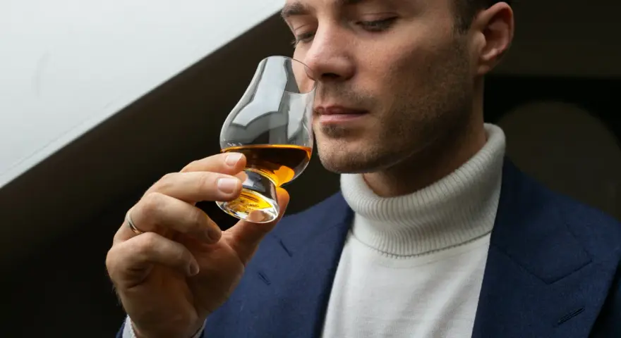 Mężczyzna w eleganckim garniturze i golfie wącha bursztynowy płyn w kieliszku. Czy to single malt?