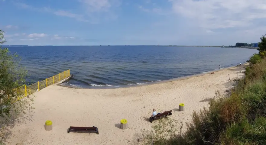 Piękna, piaszczysta plaża dla psów z łagodnym zejściem do morza. W oddali widać falochron i statki.