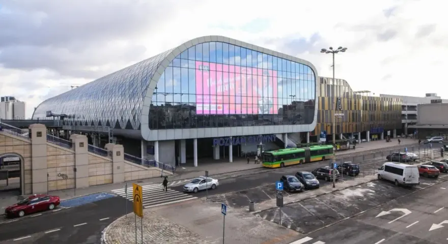 Nowoczesny Poznań Główny z charakterystyczną szklaną fasadą i ekranem. Autobusy i samochody czekają na pasażerów.