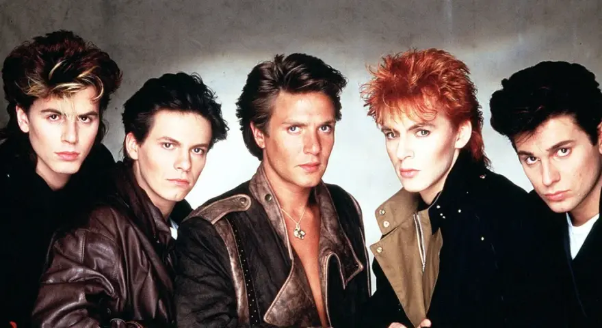 Fünf Männer mit auffälligen Frisuren und 80er-Jahre-Mode, die an ikonische Musikvideos dieser Ära erinnern.