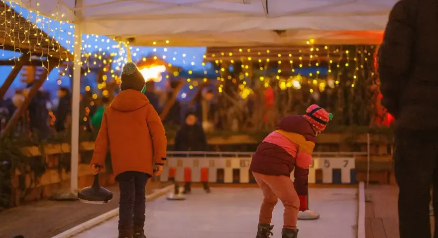 Kinder spielen Eisstockschießen auf einem Weihnachtsmarkt in Berlin. Lichterketten leuchten im Abendlicht.