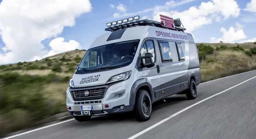 Szary kamper Ducato 4x4 Expedition z bagażnikiem dachowym i dodatkowym oświetleniem jedzie po drodze wśród wzgórz.