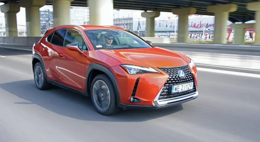 Pomarańczowy Lexus UX 250h jedzie po miejskiej drodze pod wiaduktem.