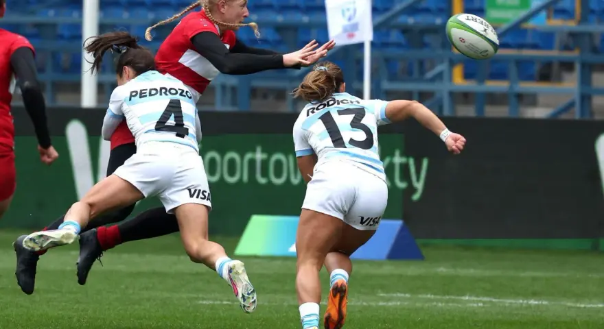 Zacięta walka o piłkę podczas rugby mistrzostwa świata. Zawodniczka w czerwonej koszulce próbuje przechwycić piłkę, podczas gdy dwie rywalki w białych strojach próbują ją zatrzymać.