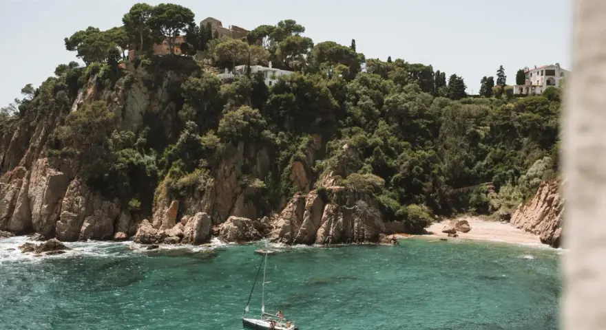 A sailboat floats in turquoise water near a rocky, tree-covered cliff with buildings. Perfect for weekend trips from Barcelona by train.
