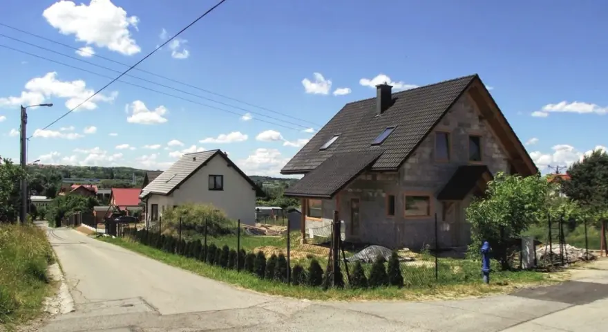 Dwa domy w budowie, jeden gotowy, na tle błękitnego nieba z chmurami. Widać drogę i zieleń. Różne kategorie gleby widoczne na poboczu.