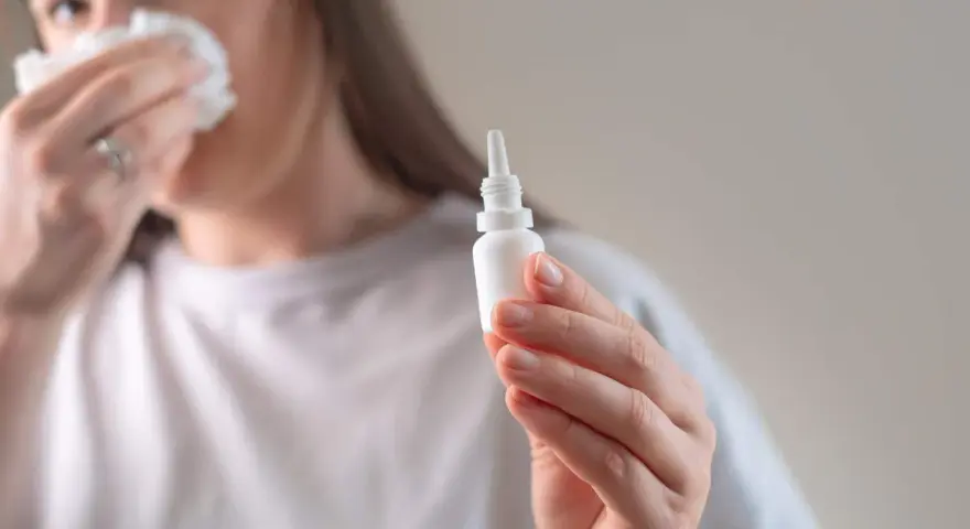 Kobieta z chusteczką przy nosie trzyma buteleczkę z kroplami do nosa, przygotowując się do plukania zatok.