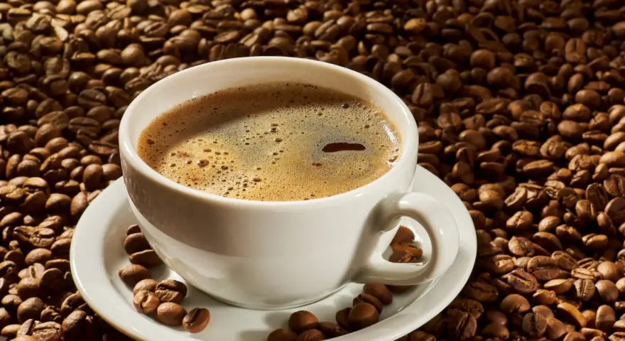 A white cup of coffee sits on a saucer surrounded by roasted coffee beans. Czy kawa rozpuszczalna podnosi ciśnienie?