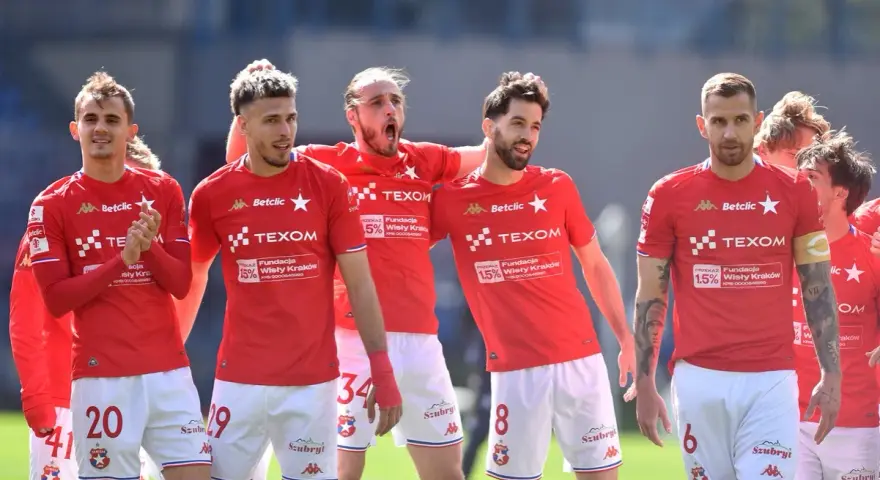 Piłkarze Wisły Kraków w czerwonych strojach, z numerami na koszulkach i spodenkach, świętują. W tle trybuny. #wisla #krakow #transfery