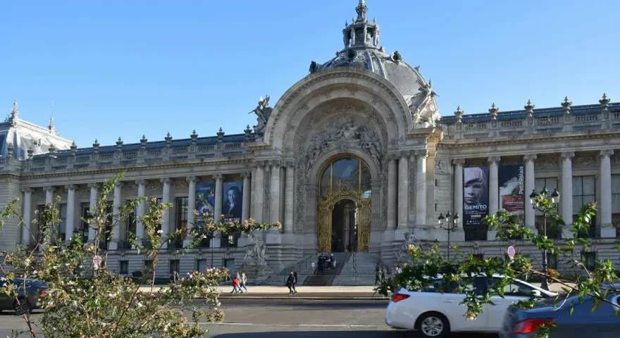 El Petit Palais, un edificio majestuoso con una cúpula y una entrada dorada, exhibe carteles de arte bajo un cielo azul.