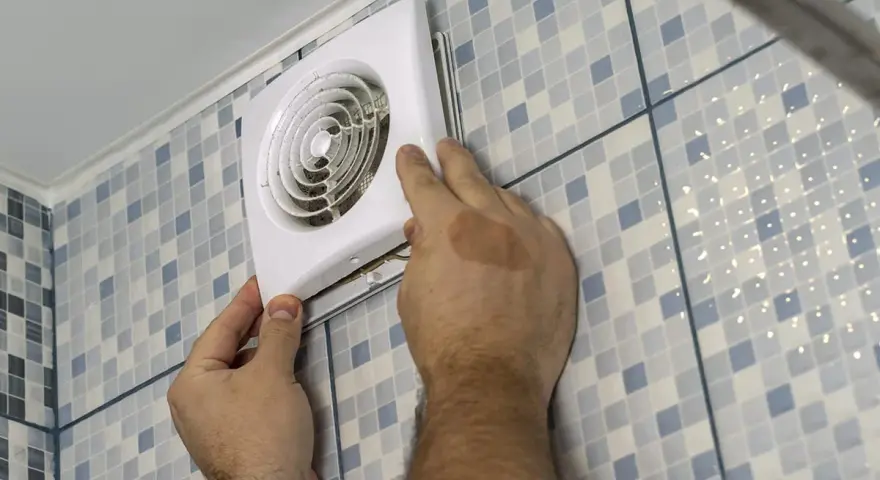 Mains d'un homme installant un ventilateur, une étape clé pour une bonne installation VMC dans une salle de bain carrelée.