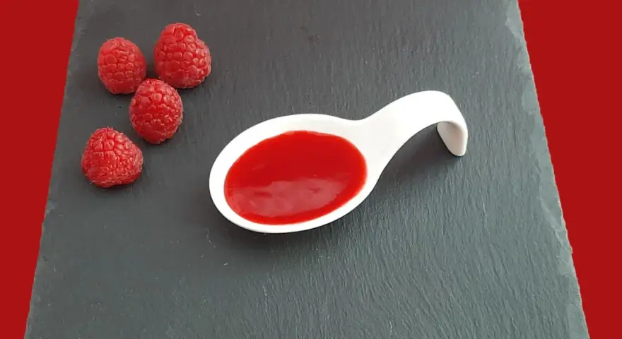 Quatre framboises fraîches à côté d'une cuillère blanche remplie de coulis de framboise, sur une ardoise.