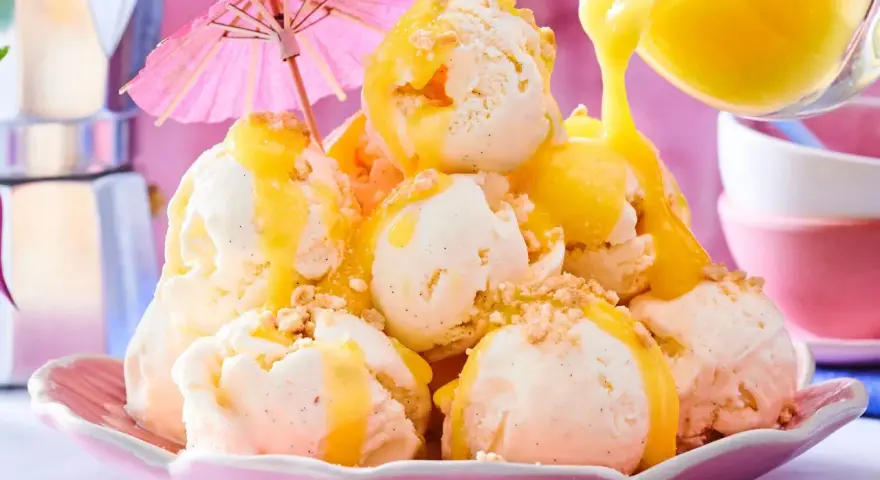 Ein Berg aus cremigem Vanilleeis, übergossen mit warmer Zitronensauce und bestreut mit Krümeln. Ein rosa Sonnenschirm sorgt für tropisches Flair.