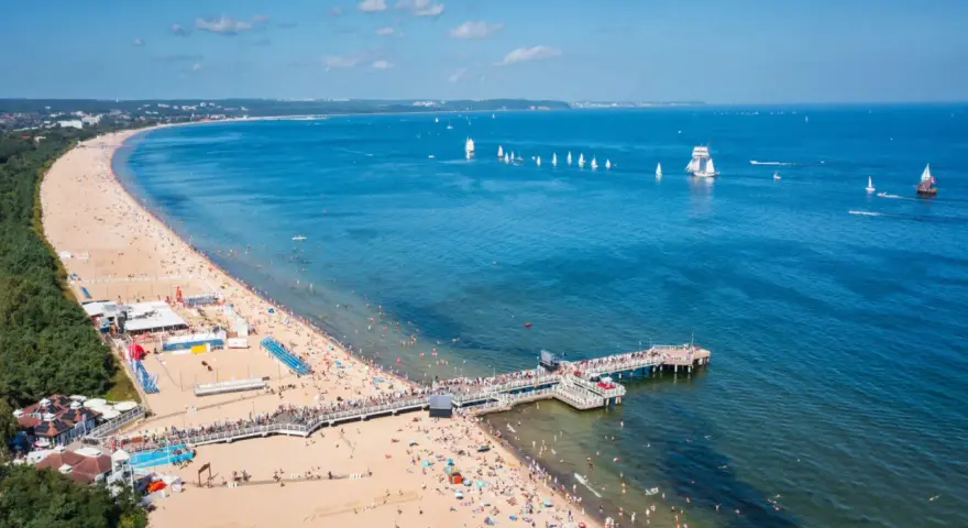 Słoneczna plaża z molo, tłumy ludzi i żaglówki na morzu – idealne miejsce na tanie wycieczki z Gdańska.