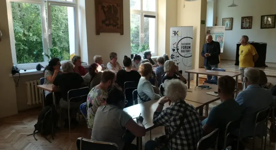 Spotkanie promujące język kaszubski. Ludzie siedzą przy stołach, słuchając prelegentów.