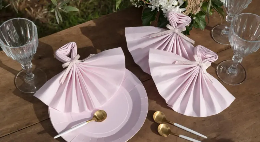 Vackert dukat bord med rosa servetter vikta som klänningar, guldfärgade skedar och glas. En bukett blommor fulländar dukningen.