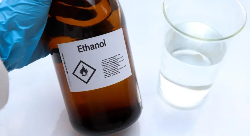Braune Glasflasche mit der Aufschrift "Ethanol" und einem Warnsymbol. Daneben ein Becherglas mit klarer Flüssigkeit. Hier gibt es 100 prozentigen Alkohol.