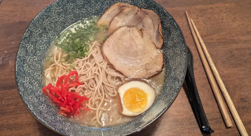 Miseczka z ramen: makaron, plastry mięsa, jajko na pół, zielona cebulka i czerwone przyprawy do ramen. Obok pałeczki.