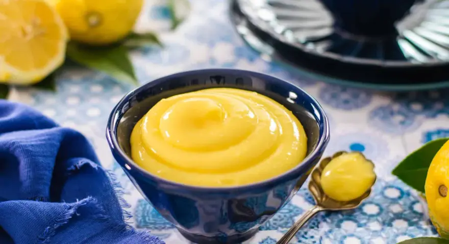 Una ciotola blu piena di vellutata crema al limone, con limoni freschi e un tovagliolo blu sullo sfondo.