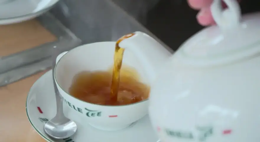 Tee mit Rum norddeutsch: Eine weiße Kanne gießt heißen Tee in eine Tasse. Ein Löffel liegt auf der Untertasse.