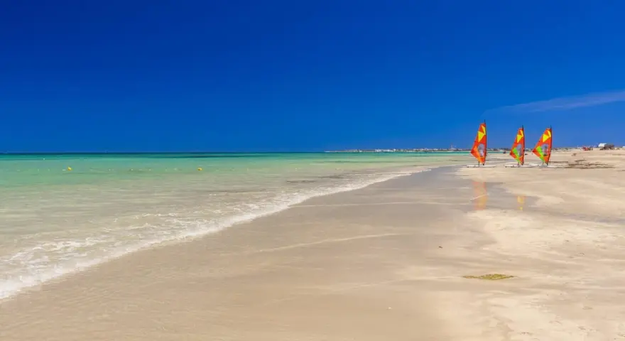 Plaża w Tunezji z trzema kolorowymi żaglami do windsurfingu na piasku i turkusową wodą.