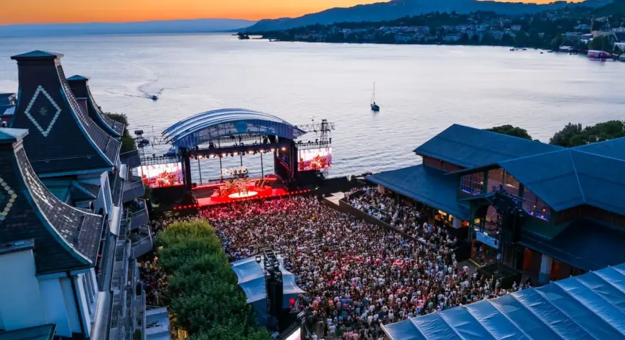 Ein großes Publikum feiert bei Sonnenuntergang ein Konzert am See. Die Bühne des Jazz Montreux Festivals ist beleuchtet, im Hintergrund der Genfersee.