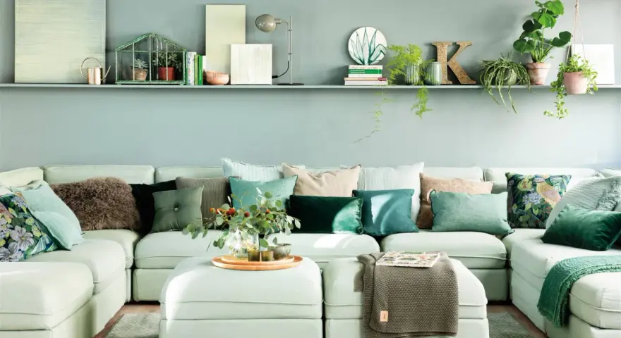 Sofá modular claro con cojines verdes y estampados, y una estantería con plantas y objetos decorativos, incluyendo una casita de cristal con **muebles pintados en verde**.