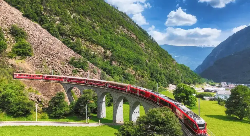 Czerwony pociąg Bernina Express przejeżdża przez kamienny wiadukt w Alpach. Sprawdź bernina express cena, by zaplanować podróż.