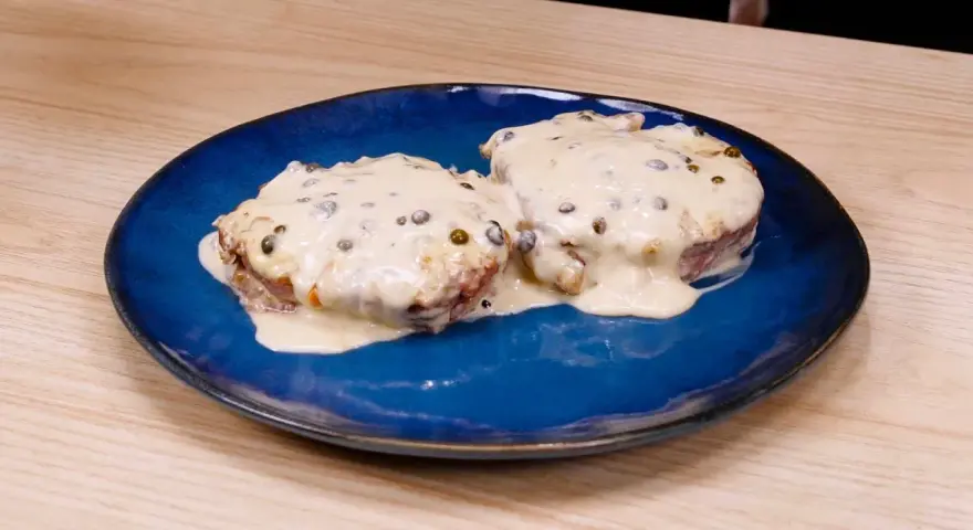 Due medaglioni di filetto al pepe verde, immersi in una cremosa salsa bianca, serviti su un piatto blu.