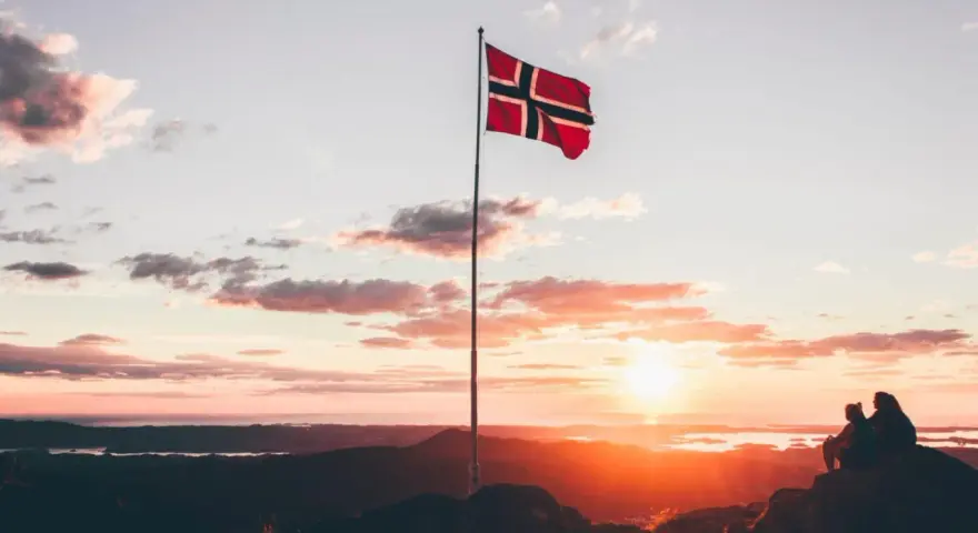 Zachód słońca w Norwegii. Dwie osoby siedzą na skale, patrząc na krajobraz. Flaga Norwegii powiewa na maszcie. Czy do Norwegii można lecieć na dowód osobisty?