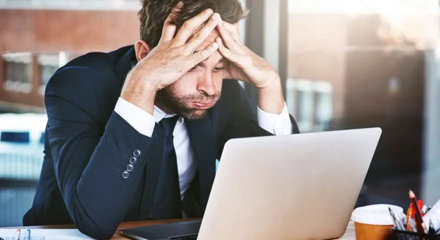 Zestresowany mężczyzna w garniturze z rękami na głowie, patrzący na laptopa. Trudna spłata długu po ogłoszeniu upadłości.