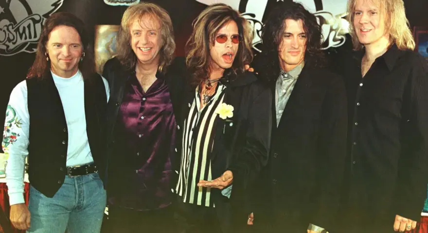 Steven Tyler mlody z zespołem Aerosmith, w charakterystycznych okularach i stroju, gotowy na scenę.