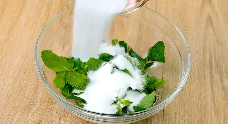 Zucchero bianco viene versato su foglie di menta fresca in una ciotola di vetro, per preparare uno sciroppo di menta fatto in casa.