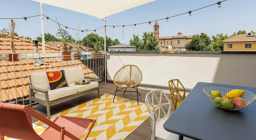 Terrazza sul tetto idee: divano, sedie colorate, tappeto giallo e bianco, tavolino con frutta, luci e vista sui tetti.